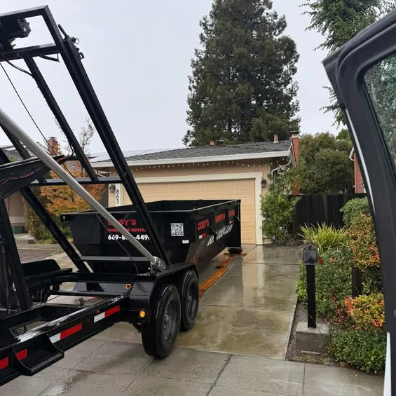 Black roll-off dumpster being delivered to a residential driveway on a rainy day, placed in front of a beige garage for a home cleanout or renovation project.