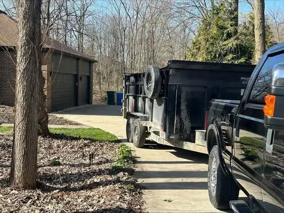 Black dump trailer parked in a wooded residential driveway next to a brick home, ready for junk removal or home renovation waste. Suitable for residential dumpster rentals in Saginaw.