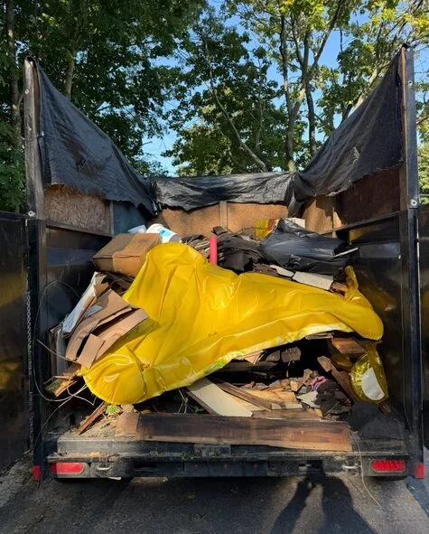Dump trailer filled with mixed household junk, cardboard, broken furniture, and a bright yellow tarp covering part of the load during a residential cleanout.