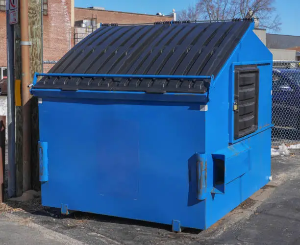 Blue front load dumpster positioned behind a commercial building for scheduled trash pickup.